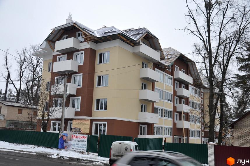 Багатоповерхова новобудова з коричневим та бежевим фасадом, білими балконами та вікнами. Навколо будинку огорожа, дерева без листя та засніжена дорога.