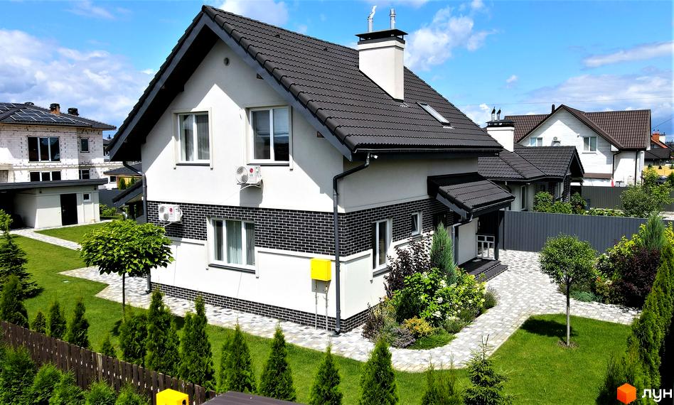 Котеджне містечко КМ Сонячний: двоповерховий будинок з темним дахом, білими стінами та декоративною цеглою. Доглянутий двір з газоном, деревами та бруківкою.