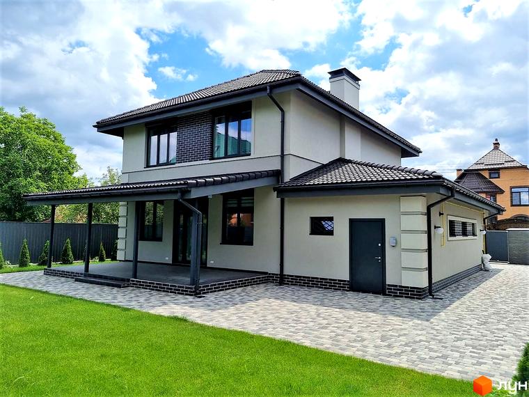 Зовнішній вигляд двоповерхового будинку з навісом та бруківкою біля котеджного містечка КМ Сонячний. Зелений газон та дерева навколо новобудови.