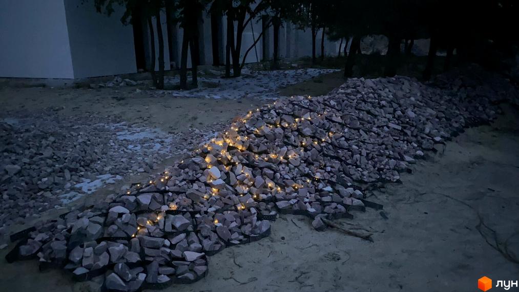 Купа щебеню з гірляндами вогнів на території забудови. Поруч видно будівлю та дерева.