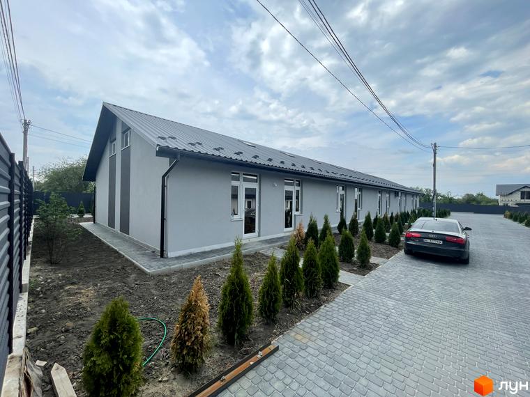 Таунхауси на Мирославській – це сучасний житловий комплекс з сірими фасадами та акуратними зеленими насадженнями. На території розташована парковка для автомобілів.