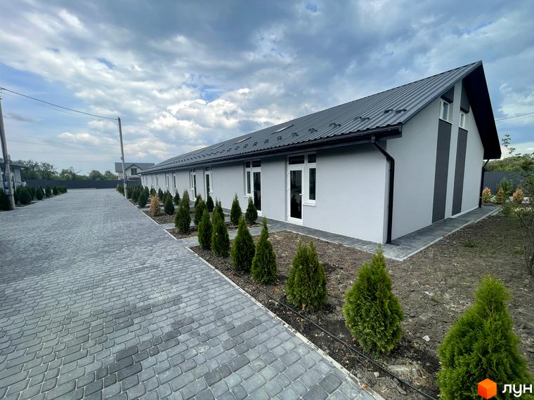 Зображення сторінки ЖК Таунхауси на Мирославській. Сіра будівля з білими вікнами та темним дахом, перед будинком викладена бруківка та висаджені туї.