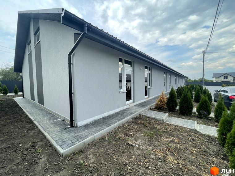 Зовнішній вигляд новобудови Таунхауси на Мирославській з сірим фасадом та бруківкою навколо. Навколо будинку висаджені молоді туї та облаштована прибудинкова територія.
