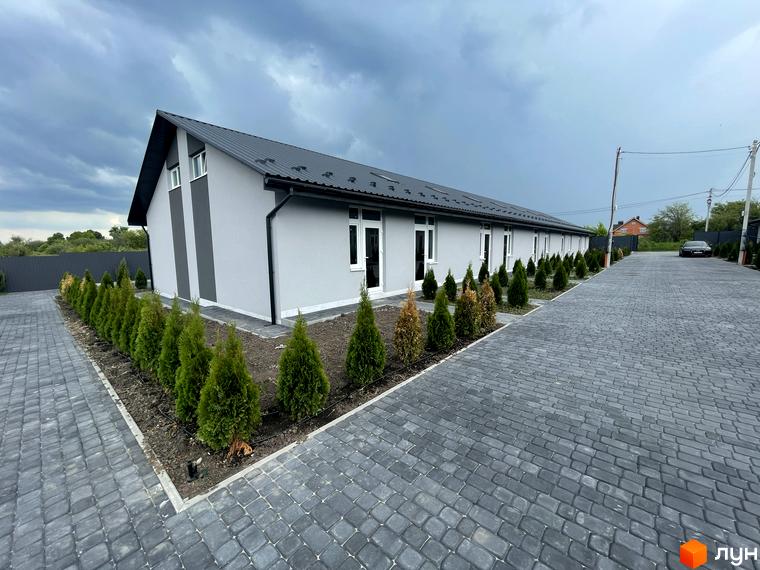 Зовнішній вигляд житлового комплексу Таунхауси на Мирославській з сірим фасадом та чорним дахом, акуратною прибудинковою територією з туями та бруківкою.