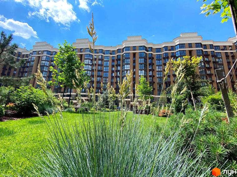 Зовнішній вигляд ЖК Sofia Nova з фасадом в коричневих тонах та великою кількістю вікон. На прибудинковій території розташований зелений газон та рослини.