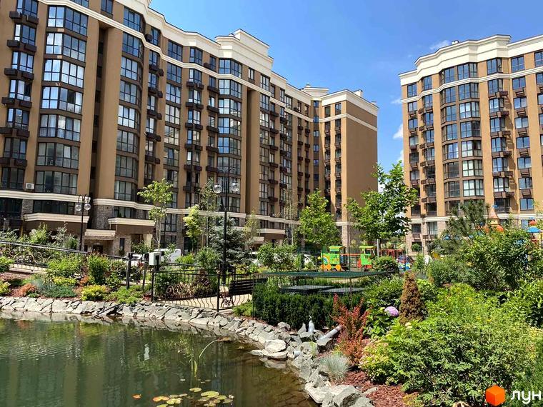 Житловий комплекс Sofia Nova з коричневими фасадами та великими вікнами. Прибудинкова територія з озелененням, водоймою та дитячим майданчиком.