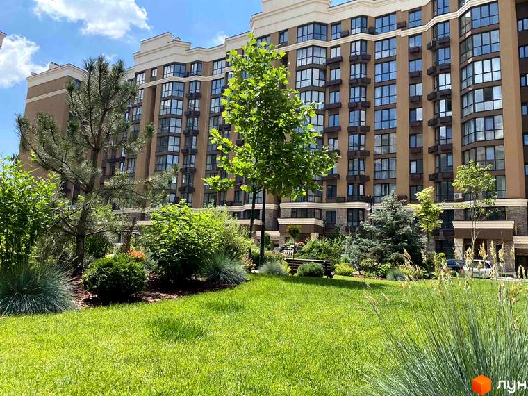 Житловий комплекс Sofia Nova з коричневим фасадом та великою кількістю вікон. Доглянута прибудинкова територія з зеленим газоном та деревами.