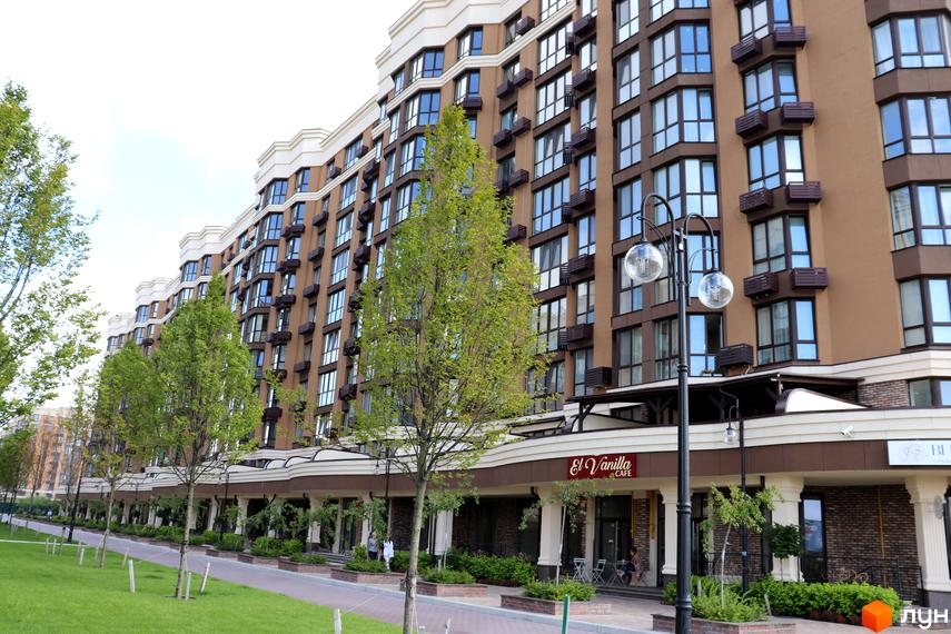 Новобудова Sofia Nova з коричневим фасадом та великою кількістю вікон. Навколо будинку зелена територія з деревами та тротуаром.