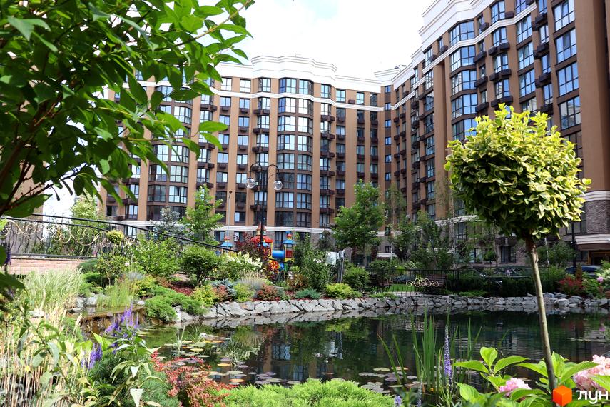 Житловий комплекс Sofia Nova з коричневими фасадами та великою кількістю вікон. На прибудинковій території розташований ставок з рослинністю та дитячий майданчик.