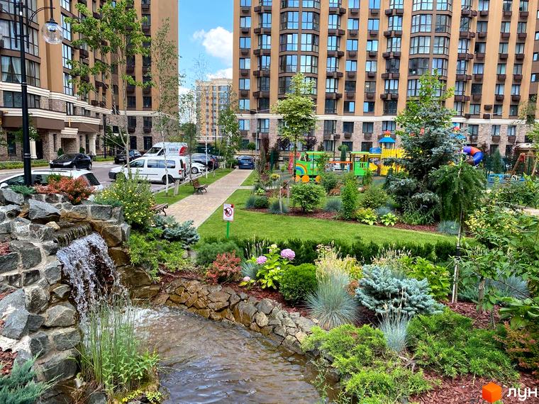 Житловий комплекс Sofia Nova: фасади будинків та доглянута прибудинкова територія з водоспадом та дитячим майданчиком. Зелені зони та зони відпочинку для мешканців.