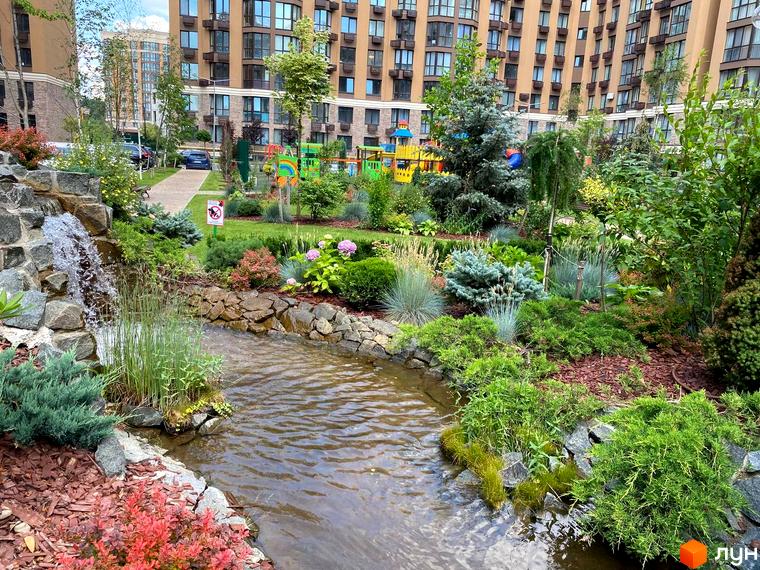 Житловий комплекс Sofia Nova: багатоповерховий будинок з фасадом з цегли та доглянутою прибудинковою територією з водоймою та зеленню. Дитячий майданчик на задньому плані.