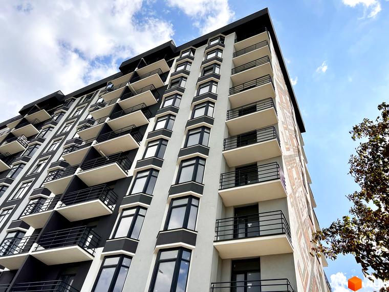 Зовнішній вигляд будинку житлового комплексу Хмельницький з численними балконами та вікнами на фоні блакитного неба. Новобудова має сіро-білу фасадну частину з декоративними елементами.