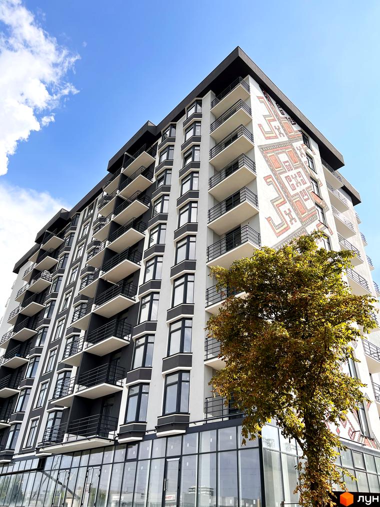 Житловий комплекс Хмельницький з фасадом, прикрашеним етнічним орнаментом, та деревом на тлі блакитного неба.