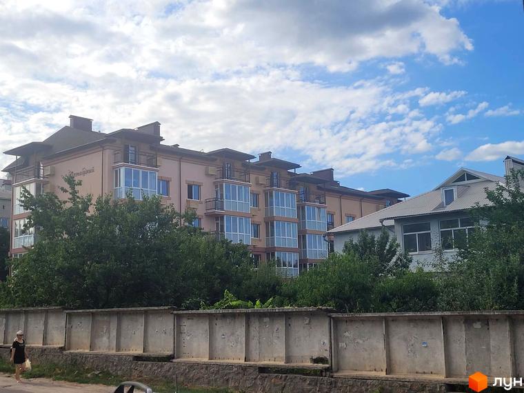 Фасад Житлового комплексу Затишний з численними заскленими балконами. Перед будинком видно зелені насадження та бетонний паркан.