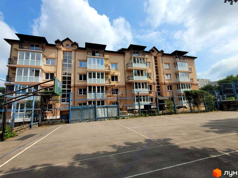 Житловий комплекс Затишний бежевого кольору з балконами та великими вікнами. Перед будинком розташований спортивний майданчик з баскетбольним кільцем та огорожею.