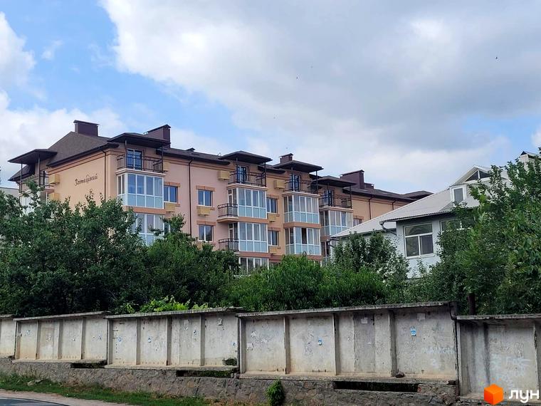 Житловий комплекс Затишний з балконами та панорамними вікнами на фоні блакитного неба. Перед будинком розташована зелена зона та бетонний паркан.