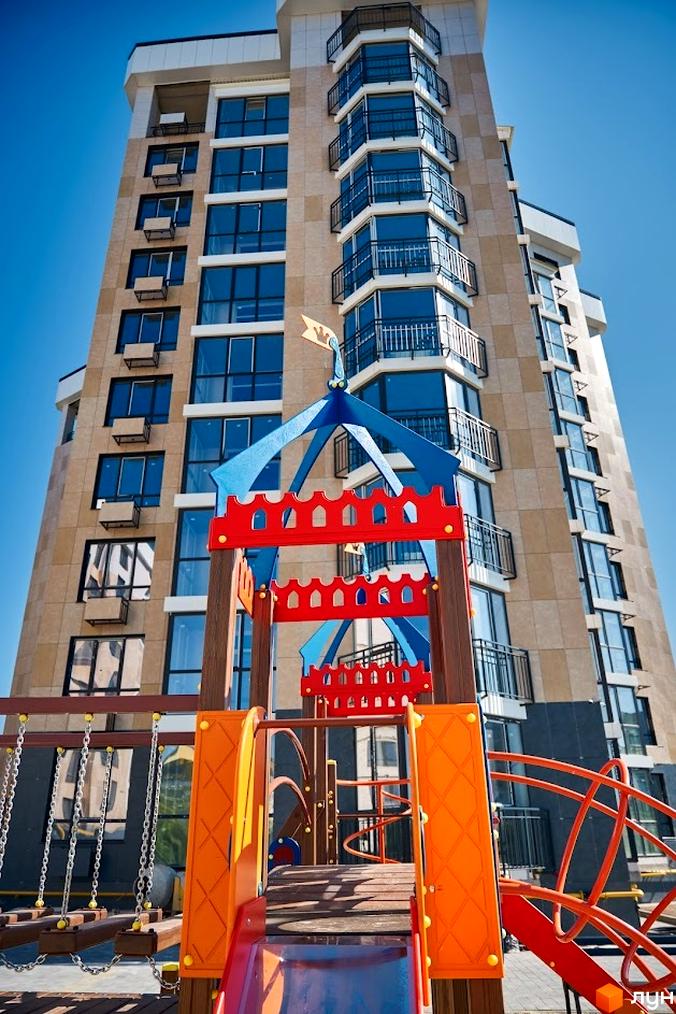 Багатоповерховий будинок Житлового комплексу Оселя Парк з бежевим фасадом та великими вікнами. На передньому плані розташований яскравий дитячий майданчик з гіркою та гойдалками.