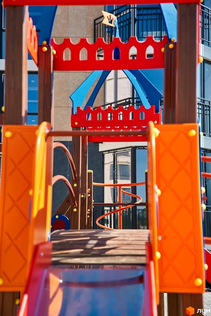 Житловий комплекс Оселя Парк з багатоквартирним будинком, що має великі вікна та балкони. На передньому плані – яскравий дитячий майданчик.