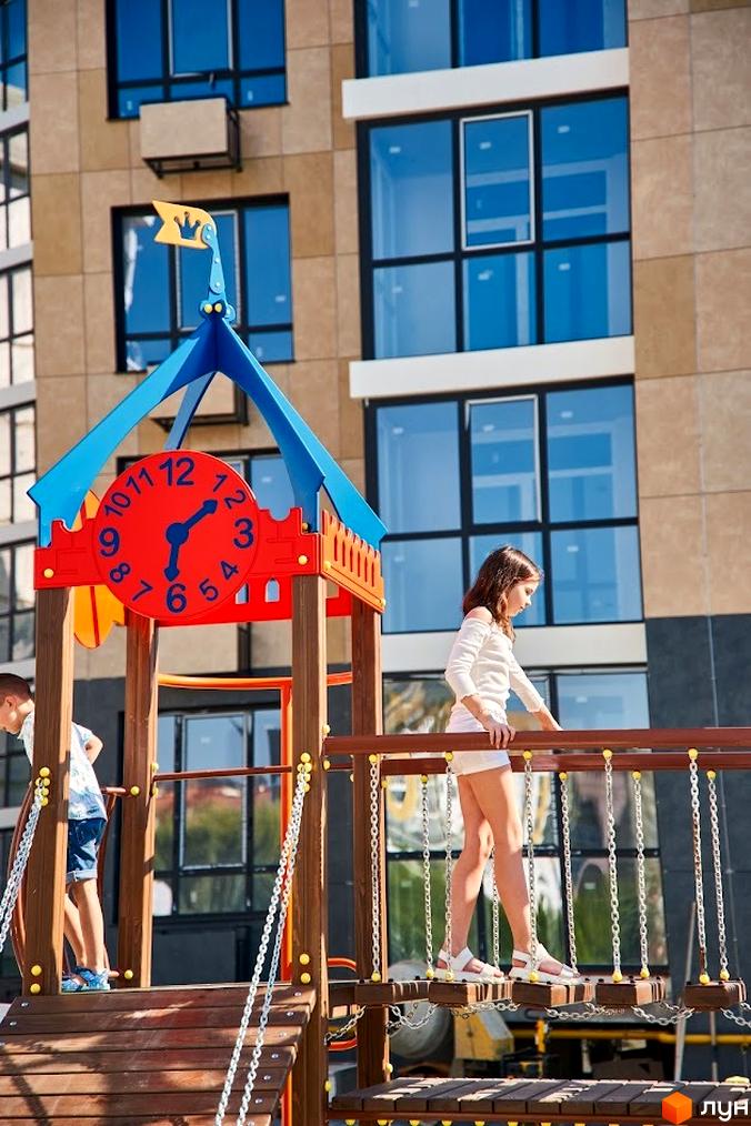 Зображення дитячого майданчика на території житлового району Оселя Парк. Вигляд фасаду будинку з великими вікнами.