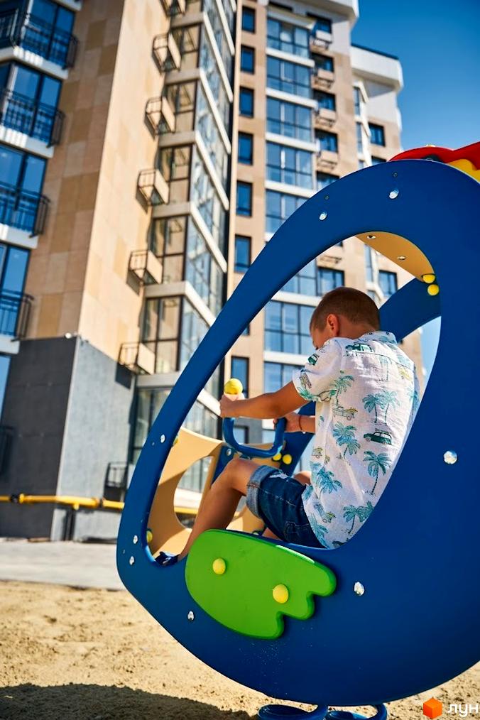 Багатоповерховий будинок Житлового комплексу Оселя Парк з великими вікнами та балконами. На передньому плані дитячий майданчик з ігровим елементом на піску.