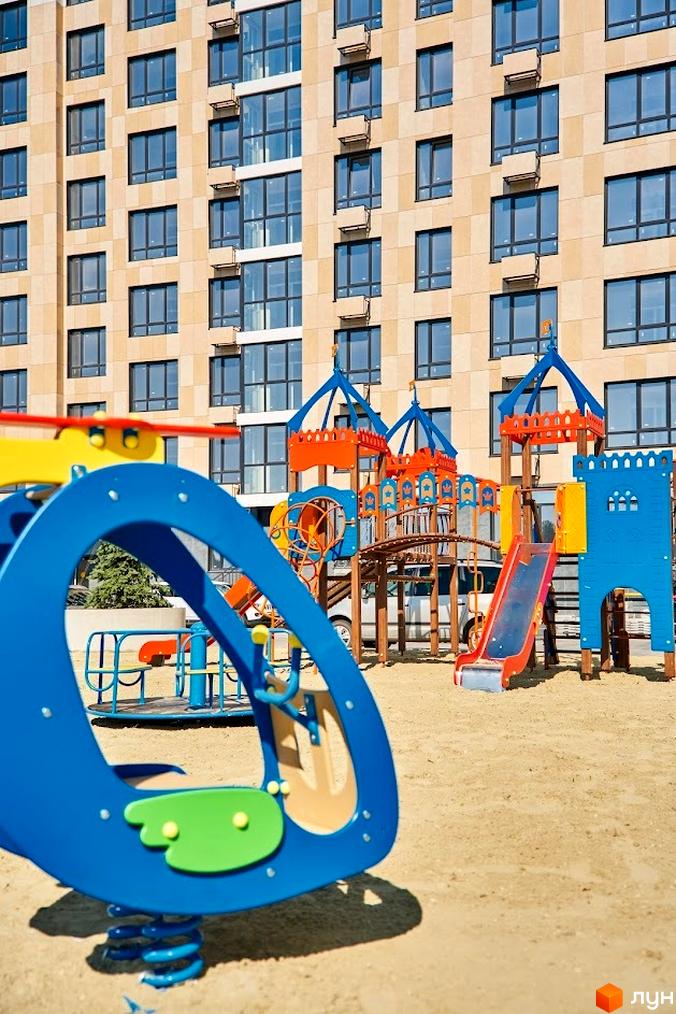 Зображення Житлового комплексу Оселя Парк з дитячим майданчиком на прибудинковій території. Високий будинок з великою кількістю вікон.