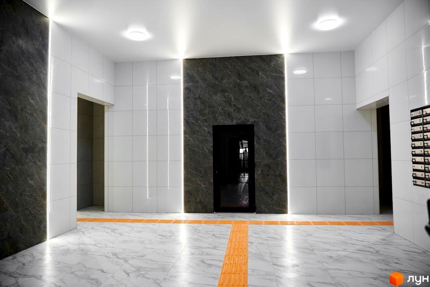 Інтер'єр вестибюлю Житлового комплексу Оселя Парк: білі стіни, темні мармурові вставки, лінійне освітлення та тактильна плитка на підлозі.