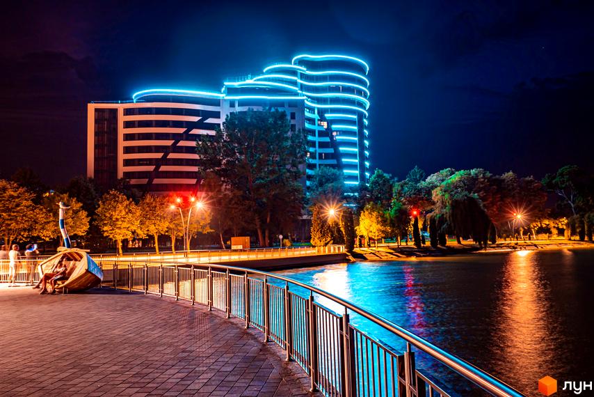 Житловий комплекс Parus з підсвіткою вночі. На передньому плані алея з лавкою у вигляді човна та водойма.