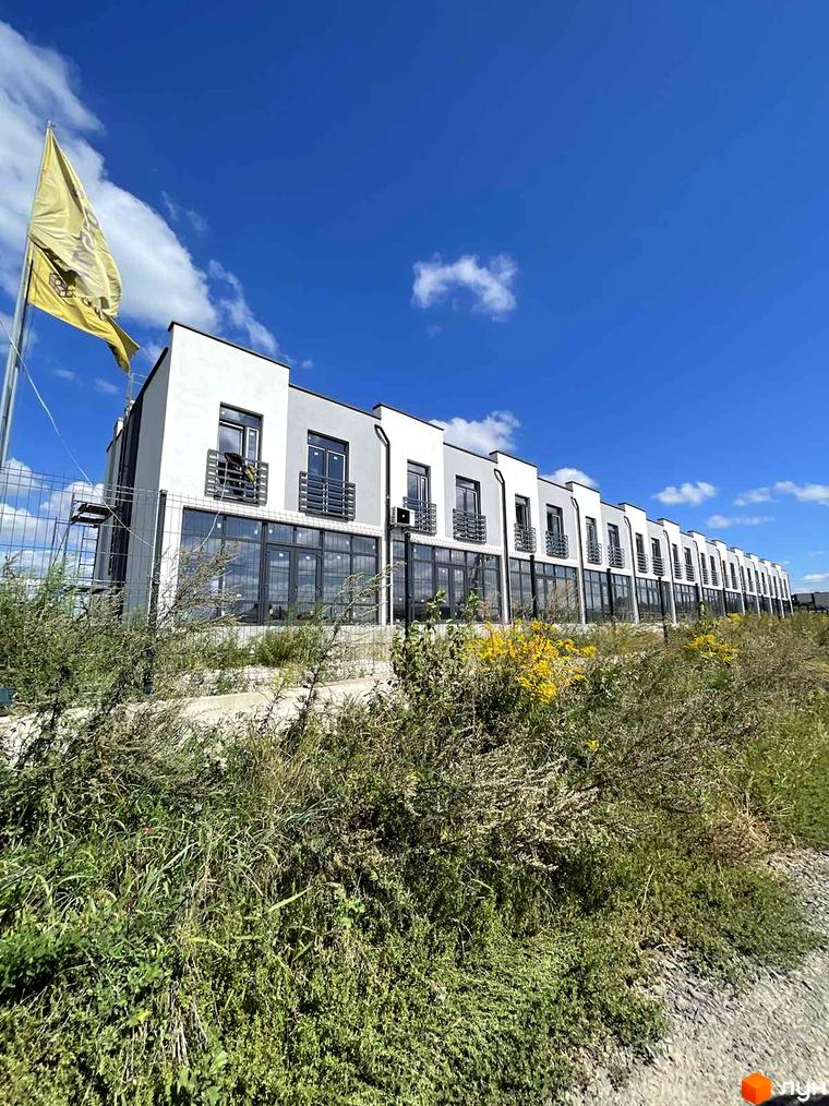 Таунхауси Green Home: ряд білих таунхаусів з темними вікнами та балконами на тлі блакитного неба, оточених зеленою рослинністю.