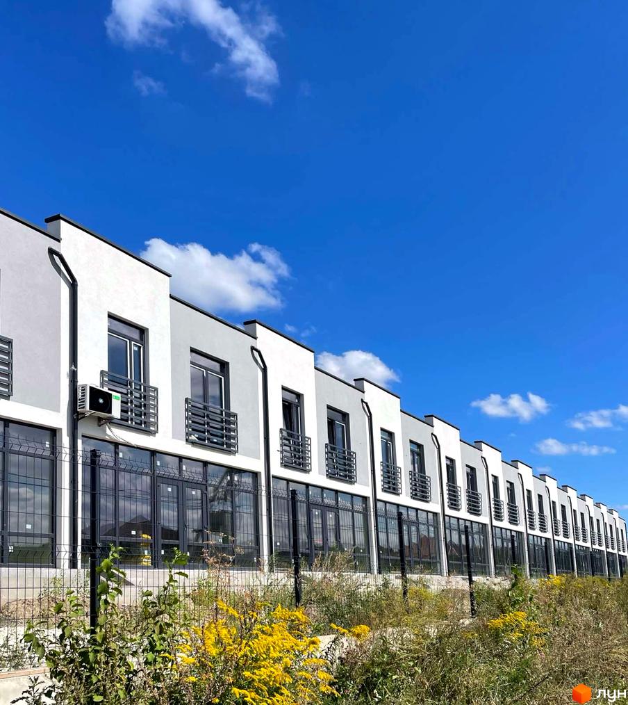 Ряд білих таунхаусів з темними вікнами та балконами на тлі блакитного неба. Перед будинками зелень та жовті квіти.