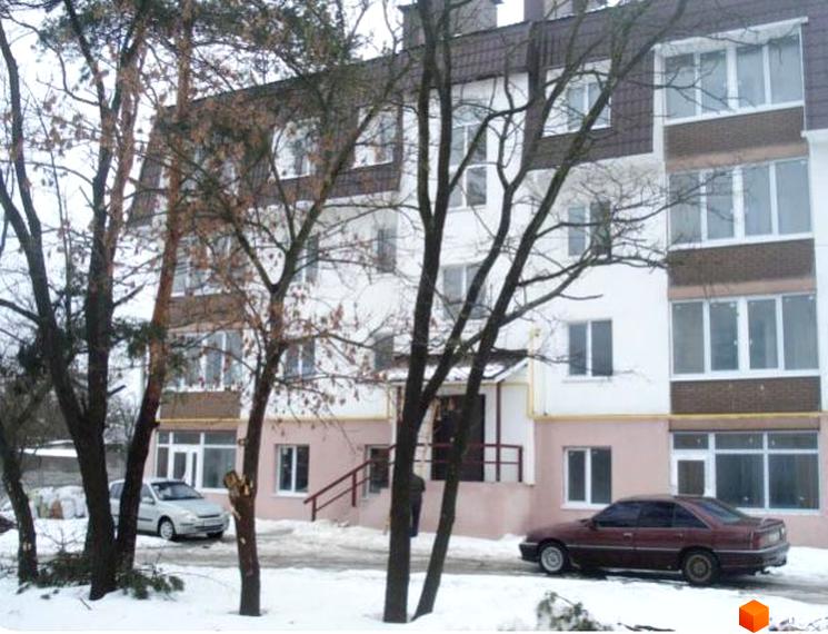 Багатоповерховий житловий будинок білого кольору з рожевими акцентами та коричневим дахом. Навколо будинку засніжена територія з деревами та припаркованими автомобілями.