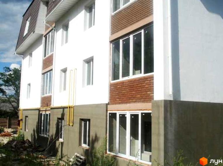 Зовнішній вигляд будинку на вул. Вериківського, 23 з білими стінами та коричневою обробкою. Прибудинкова територія з зеленими насадженнями та будівельними матеріалами.