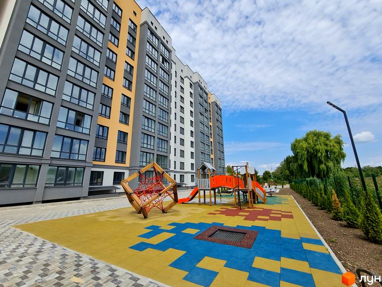 Фасад Житлового комплексу Дім виконаний у сірих, жовтих та білих тонах. Поруч знаходиться дитячий майданчик з різнокольоровим покриттям.
