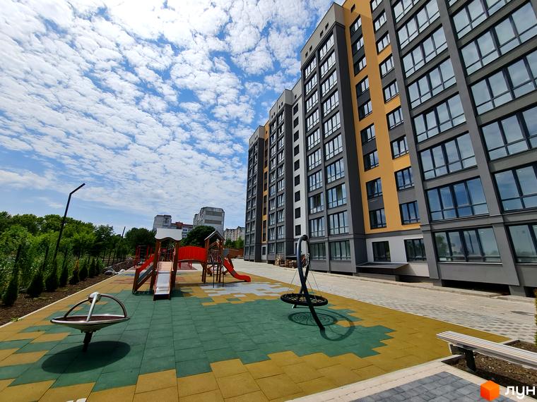 Багатоповерховий будинок Житлового комплексу Дім має сіро-жовтий фасад. Поруч облаштований дитячий майданчик з ігровим обладнанням.