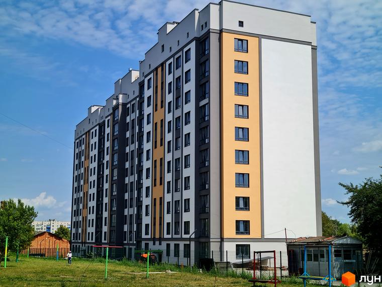 Багатоповерховий будинок Житловий комплекс Дім з білими, сірими та помаранчевими фасадами. Перед будинком зелений газон та дитячий майданчик.