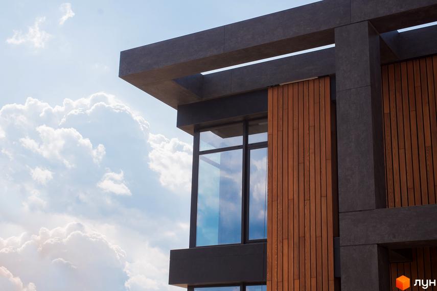 Фасад будинку Котеджного містечка 9 ЕЛЕМЕНТІВ з темними панелями та вертикальними дерев'яними елементами. Великі вікна відбивають небо з хмарами.