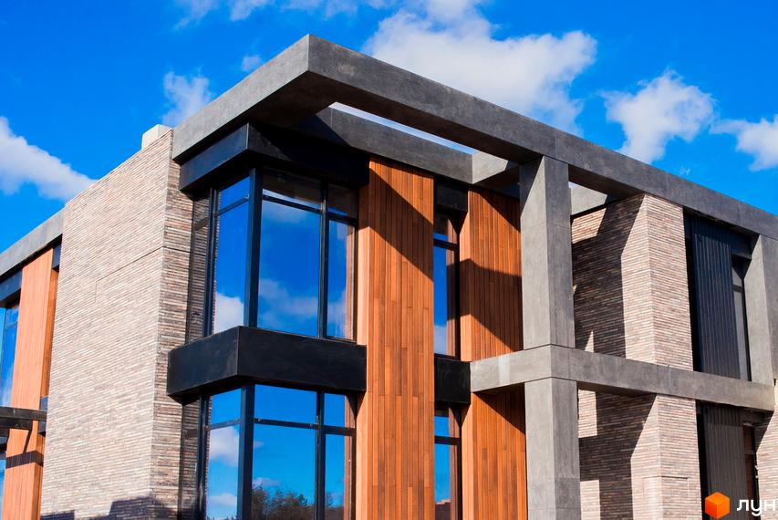 Котеджне містечко КМ 9 ЕЛЕМЕНТІВ. Зовнішній вигляд будинку з цегляними та дерев'яними елементами, великими вікнами та бетонними конструкціями на тлі блакитного неба.