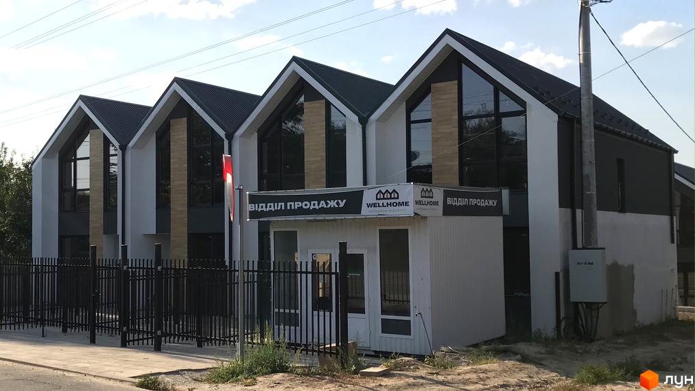 Зовнішній вигляд таунхаусів Well Home з чорним дахом та великими вікнами. На передньому плані огорожа та невелика будівля відділу продажу.