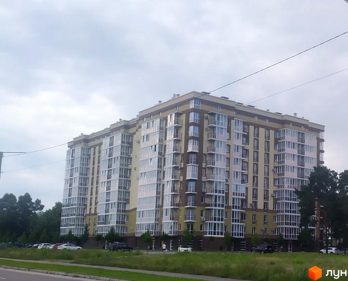 Житловий комплекс Oliva: багатоповерховий будинок світло-жовтого та коричневого кольорів з балконами. Поруч зелена зона, дорога та авто.