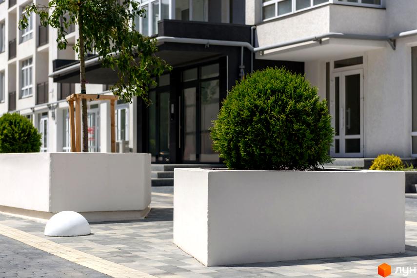 Зображення житлового комплексу KromaxBud з великими білими клумбами перед входом. На задньому плані видно фасад будівлі з великими вікнами.