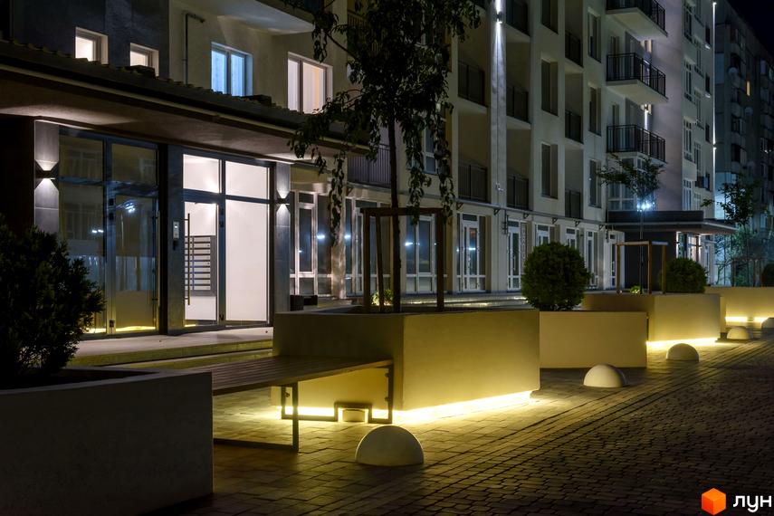 Новобудова ЖК KromaxBud з підсвіченою прибудинковою територією та лавкою. Сучасний дизайн фасаду та освітлення території.