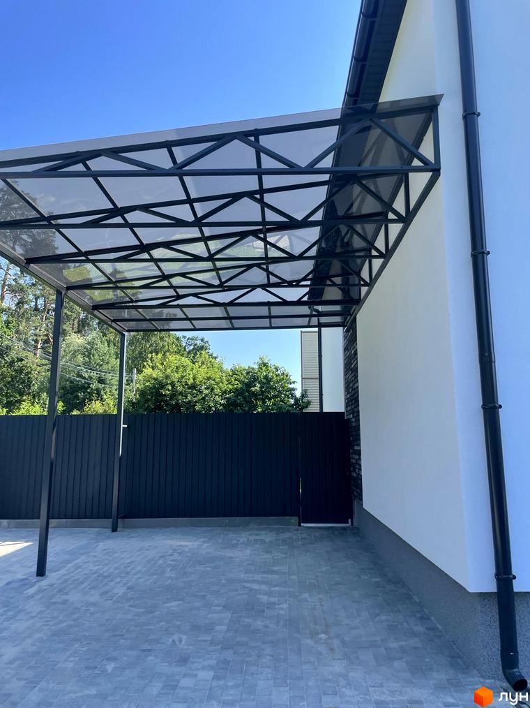 Сучасний навіс над бруківкою біля будинку з білою стіною та чорною водостічною трубою. Навколо зелені дерева та паркан.