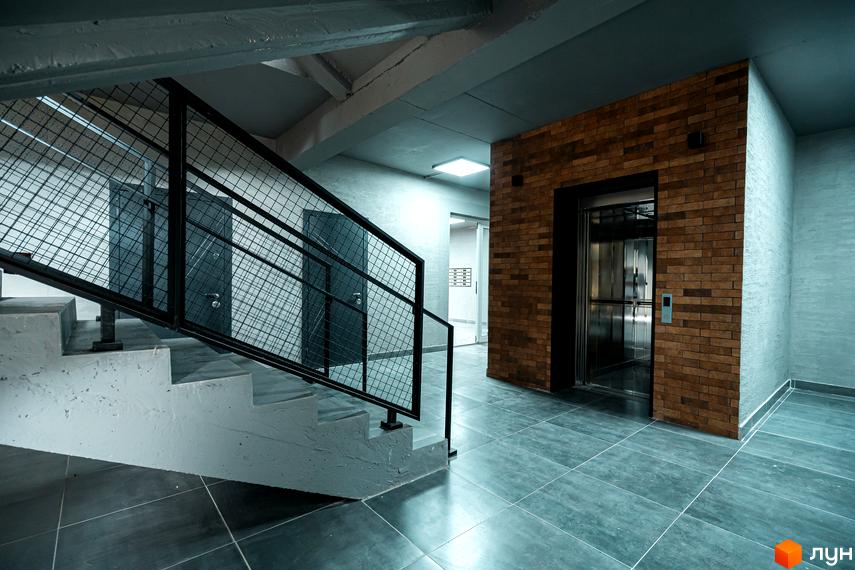 Інтер'єр сходової клітки з ліфтом у житловому комплексі Клубне містечко 12. Цегляна стіна біля ліфта та сіра підлога.
