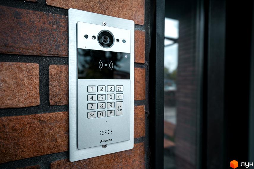 Сучасний домофон з кодовою панеллю та камерою на цегляній стіні ЖК Клубне містечко 12. Поруч розташовані елементи фасаду будівлі та вхідна група.