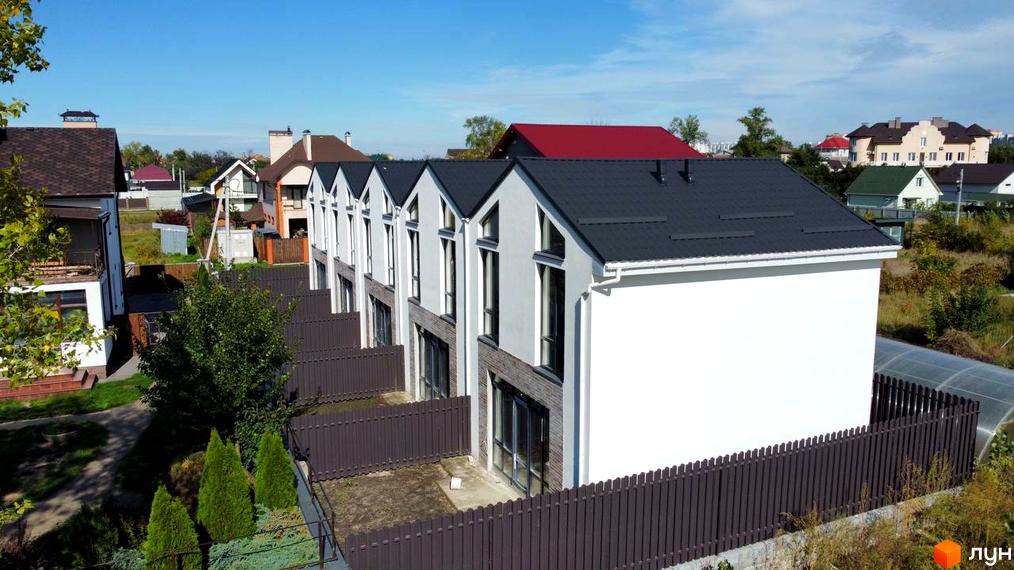 Таунхауси West Homes з темними дахами та білими фасадами, огороджені коричневим парканом. На прибудинковій території ростуть туї та зелені кущі.