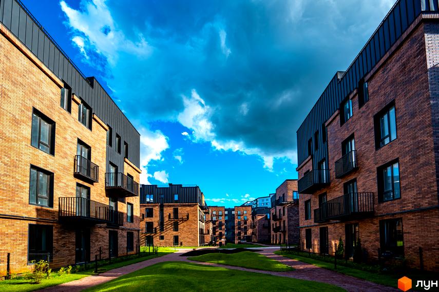 Зовнішній вигляд новобудови ЖК Клубне містечко 12 з цегляним фасадом та чорними елементами. Навколо будинку доглянута прибудинкова територія з газоном та доріжками.