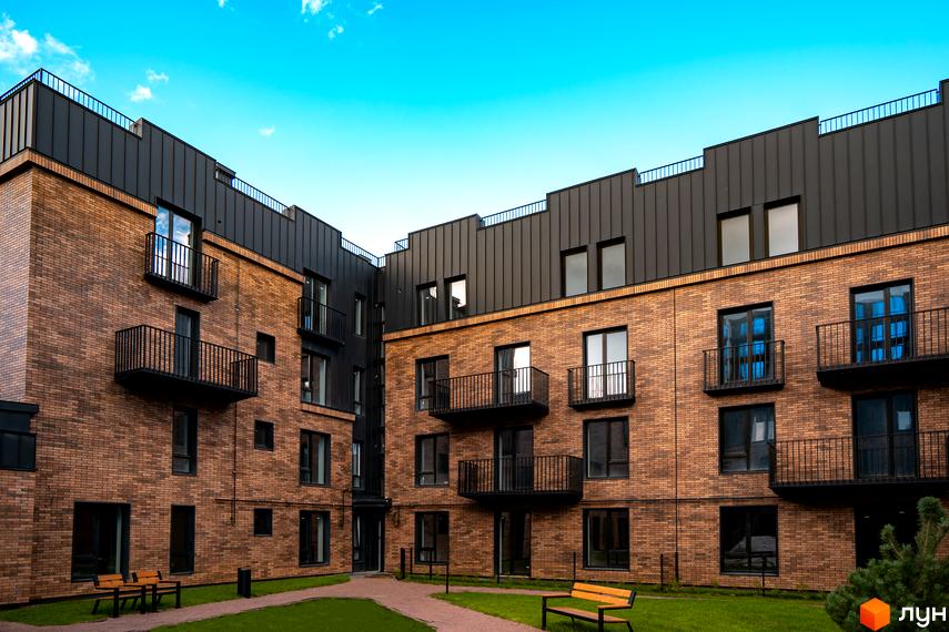 Зовнішній вигляд клубного містечка 12 з цегляним фасадом та чорним дахом. Прибудинкова територія з газоном та лавками.