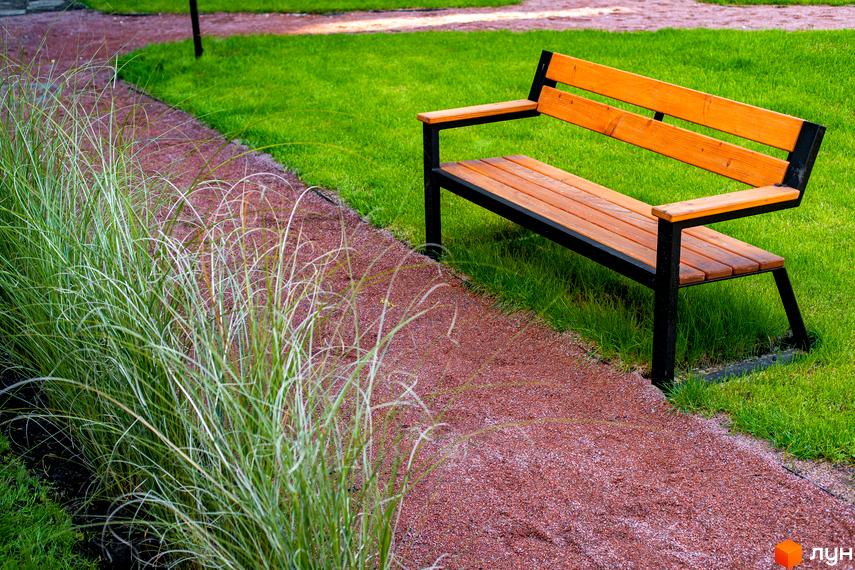 Зовнішній вигляд лавиці в парку біля доріжки з червоної крихти та зеленої трави. Клубне містечко 12 - це затишне місце для відпочинку на природі.