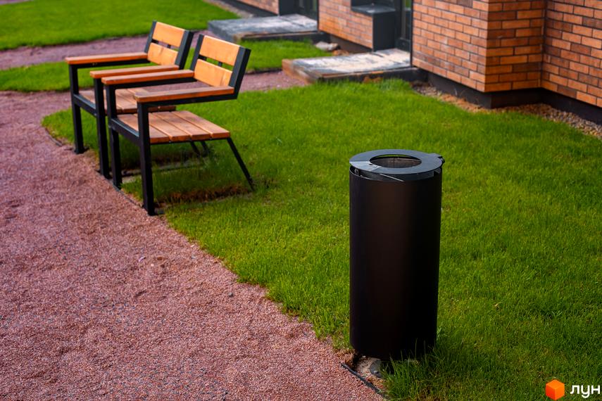 Два дерев'яні лавки з металевим каркасом на фоні зеленої галявини та цегляної будівлі. Поруч розташована чорна кругла урна.