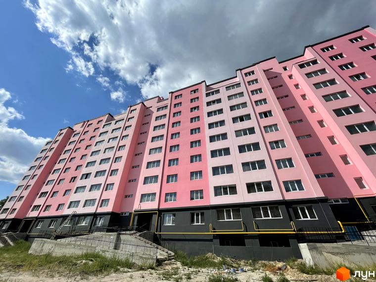 Фасад житлового комплексу Світанок у рожевих та світло-рожевих тонах з великою кількістю вікон. Біля будинку розташовані сходи та зелені насадження.