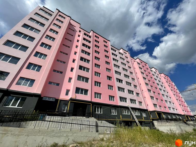 Зовнішній вигляд новобудови ЖК Світанок. Багатоповерховий будинок з рожевим фасадом та великою кількістю вікон. Навколо будинку облаштована прибудинкова територія з огорожею та зеленими насадженнями.